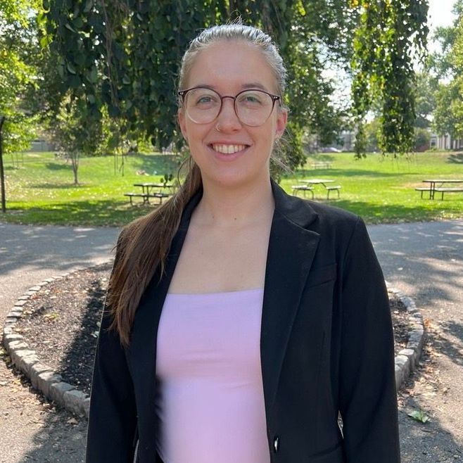 A woman wearing glasses and a black jacket is standing in a park.