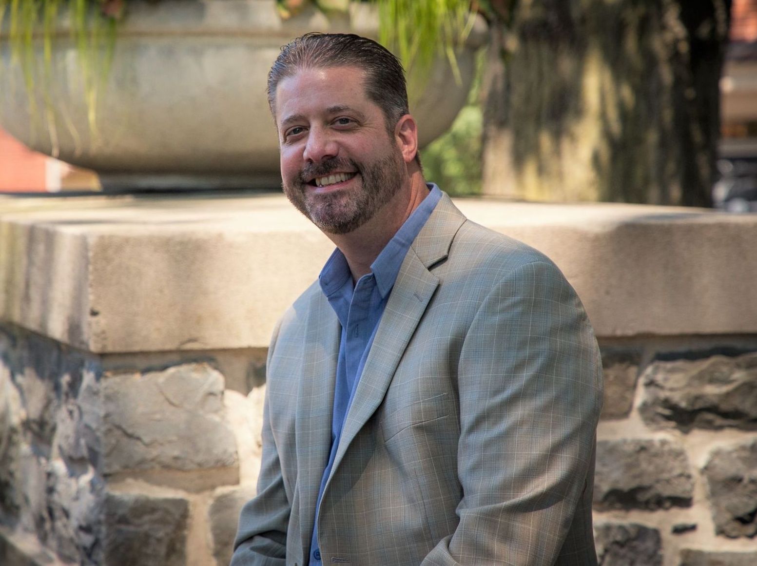 A man in a suit and blue shirt is sitting on a stone wall.