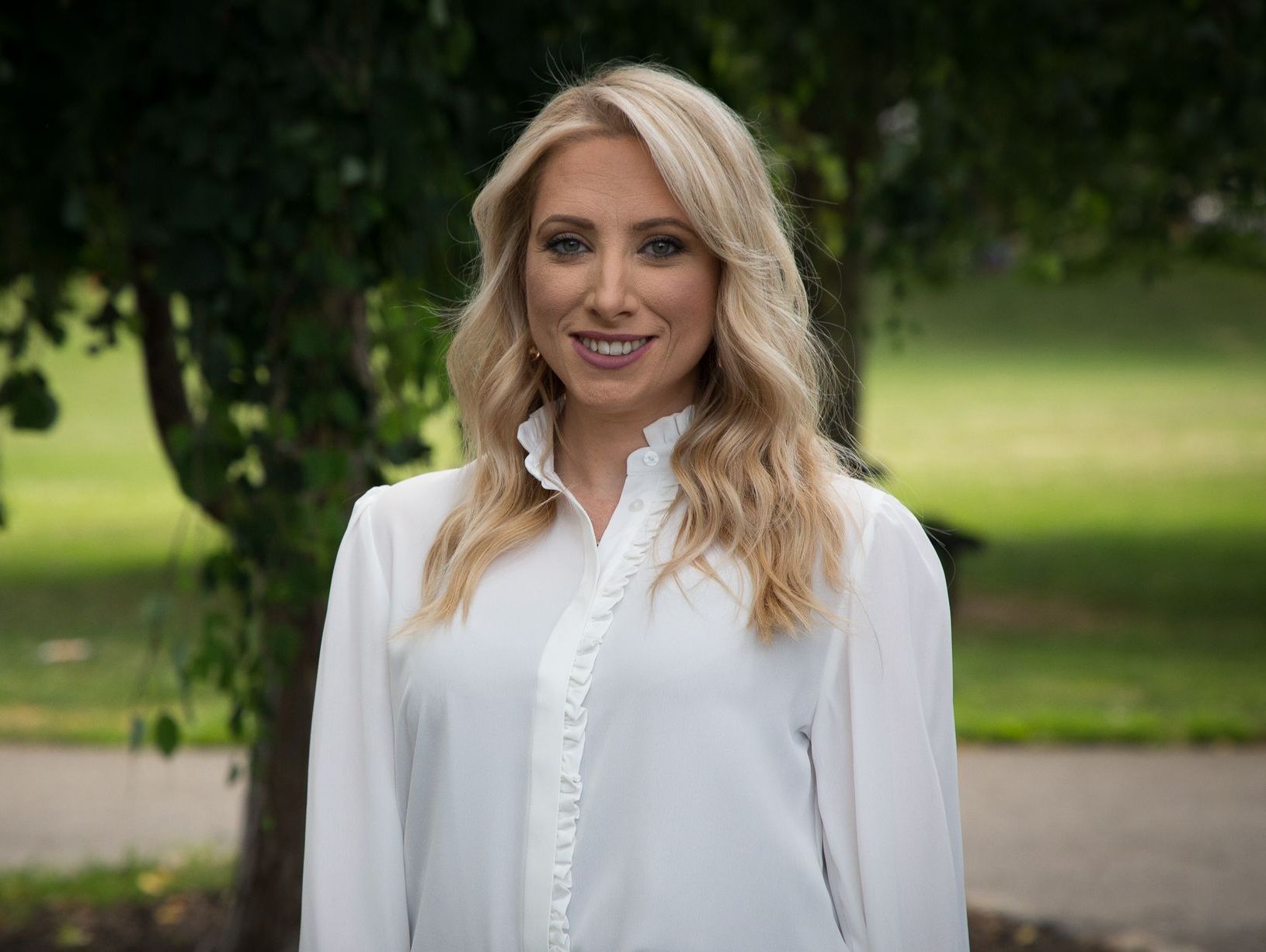 A woman in a white shirt is standing in front of a tree.