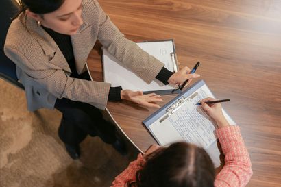 A woman is sitting at a table talking to another woman who is writing on a clipboard.