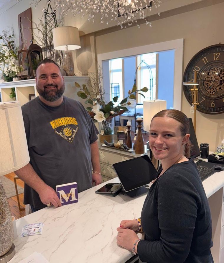 Two people smiling behind a marble counter in a bright shop, with flowers, a laptop, and wall decor.