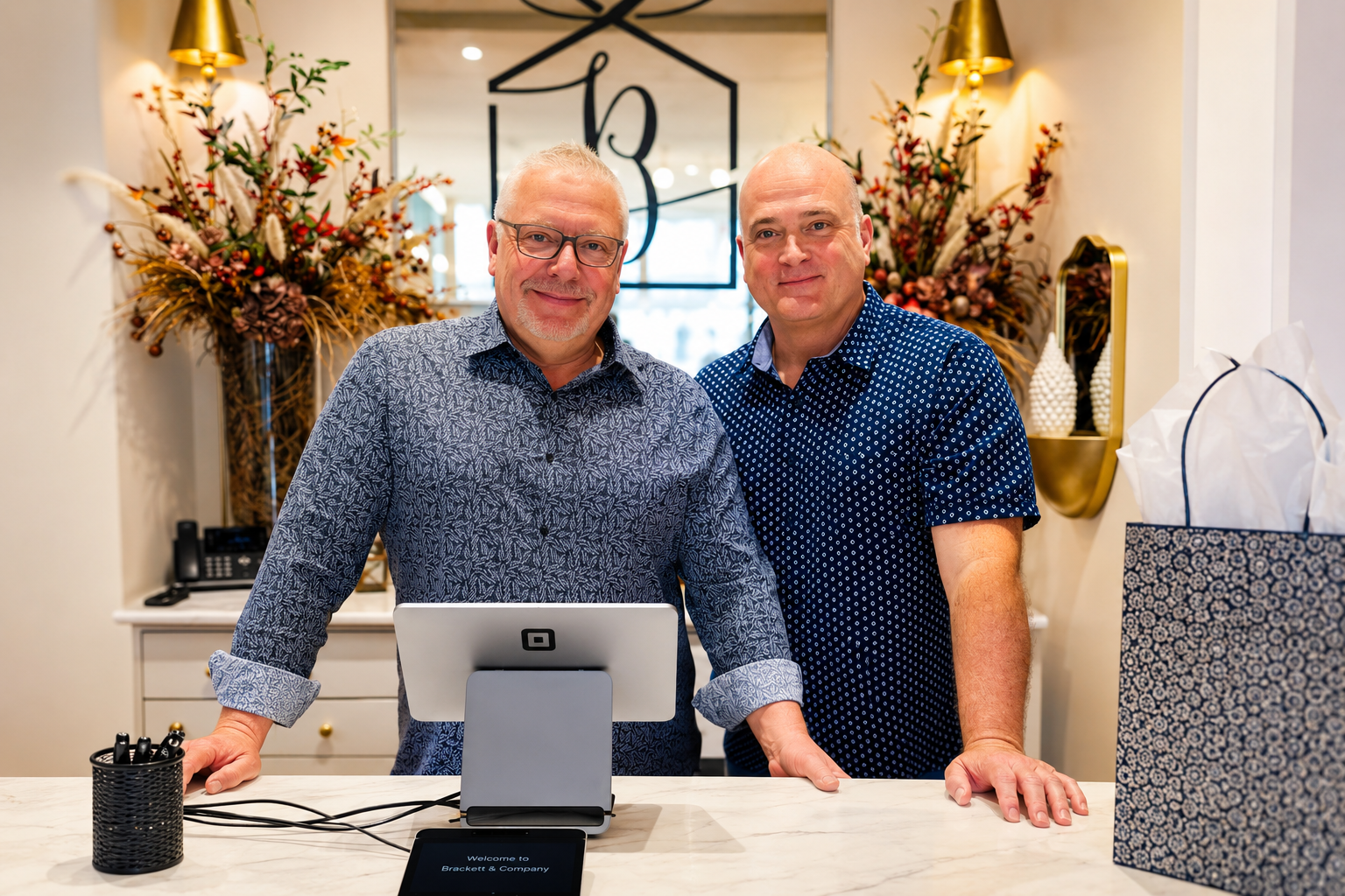 Two men stand behind a counter in a bright boutique, with flowers and gift bags in the background.