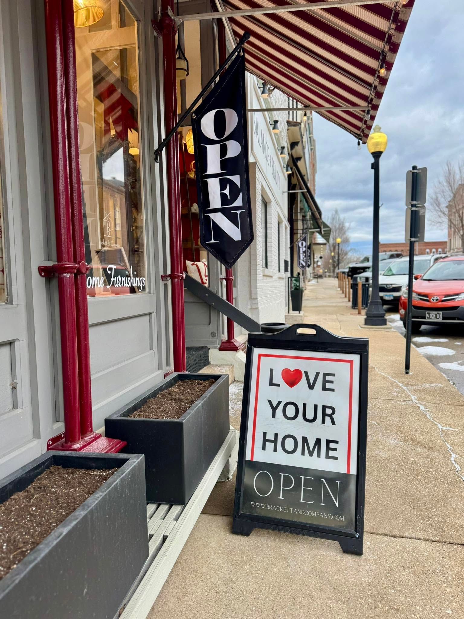 Sidewalk storefront with hanging “OPEN” sign and sandwich board reading “WE ♥ YOUR HOME OPEN.”