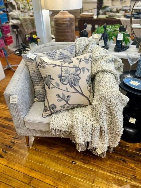 Patterned armchair with floral pillow and throw blanket in a home decor store, on a wooden floor.
