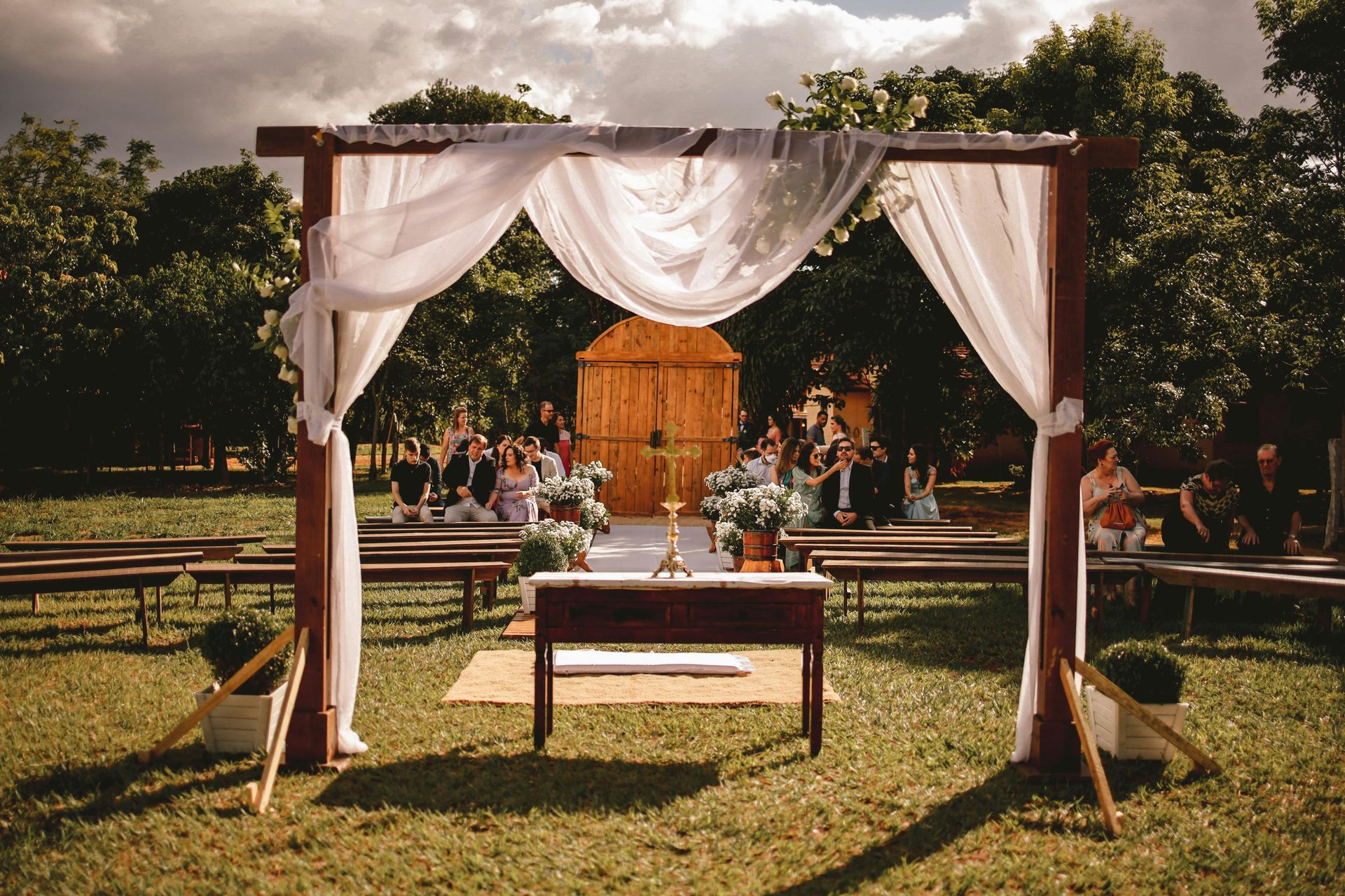 A wedding ceremony is taking place in a field with a wooden archway.