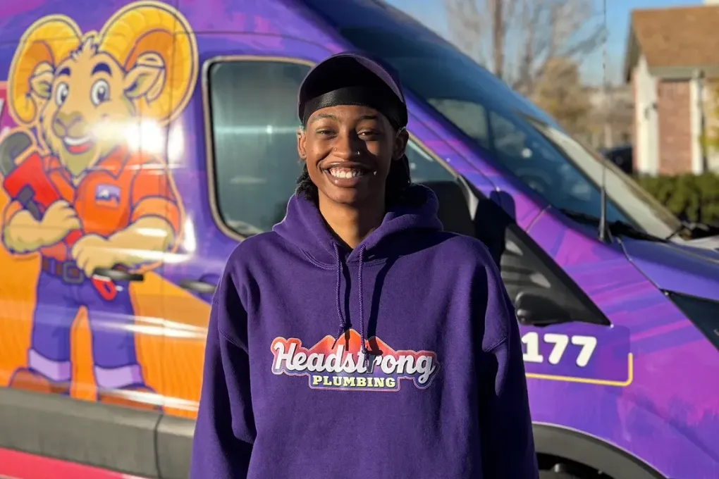 Smiling person in a purple hoodie standing beside a purple van with a cartoon mascot