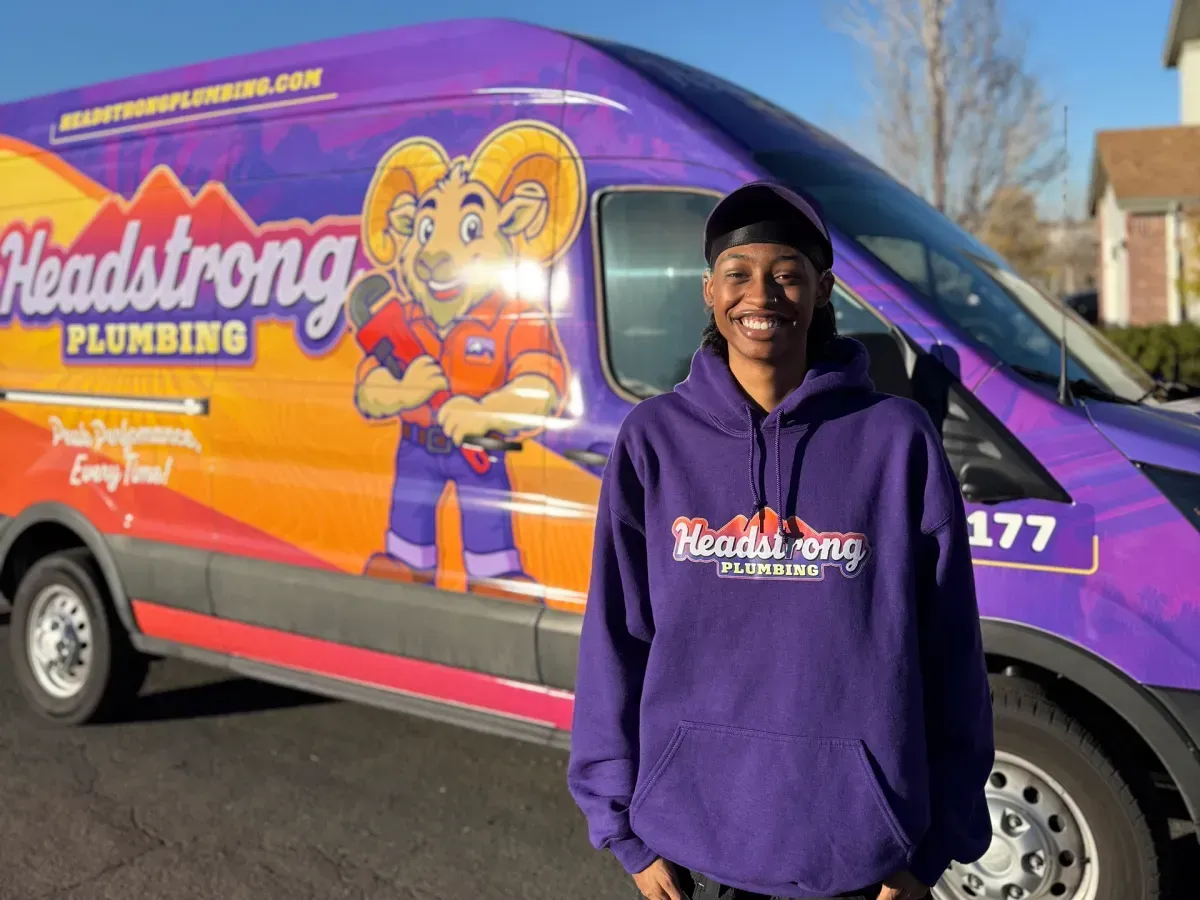 Person in purple hoodie standing beside a purple-and-orange plumbing van with cartoon mascot and logo