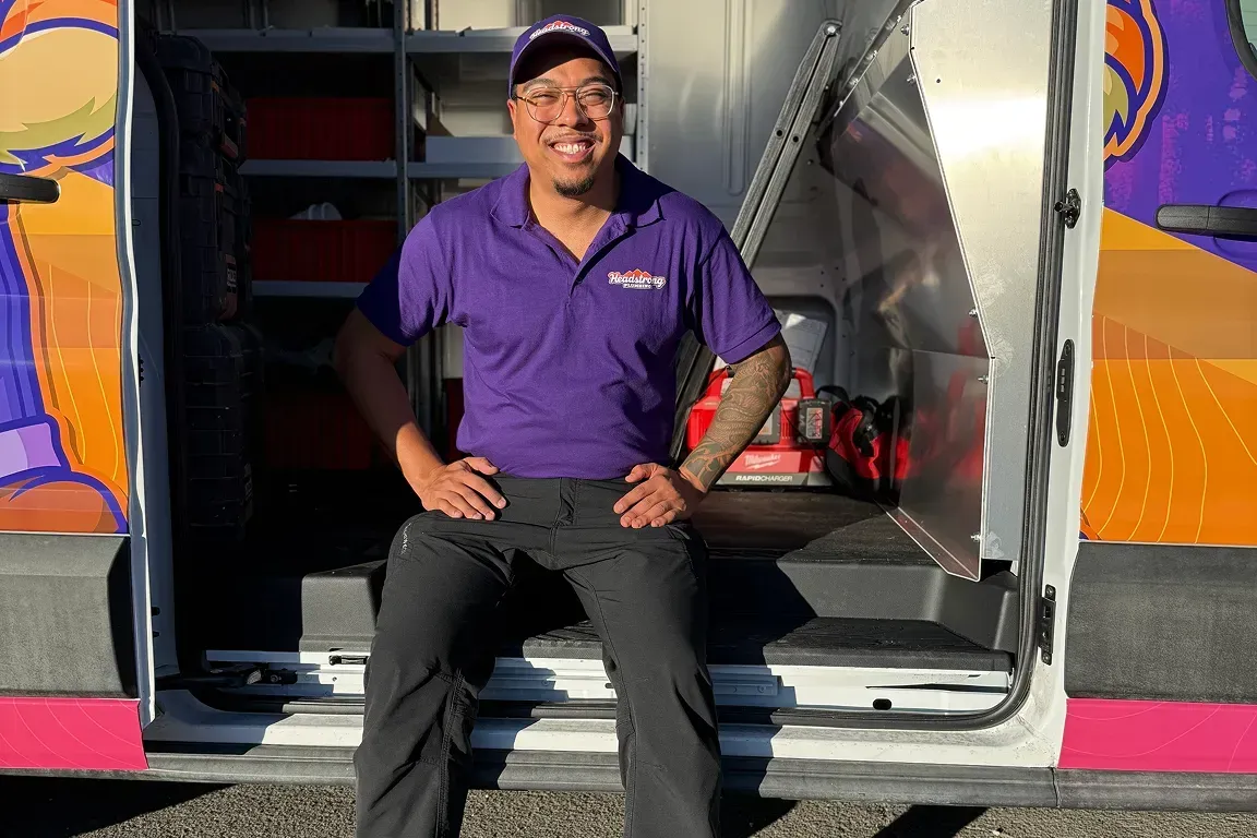 Man sitting in a van doorway, smiling in a purple shirt and cap, with a colorful vehicle exterior.