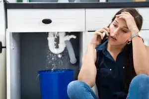 Plumbing Repairs Highlands Ranch CO Woman on phone beside leaking sink, looking stressed, with water dripping into a blue bucket