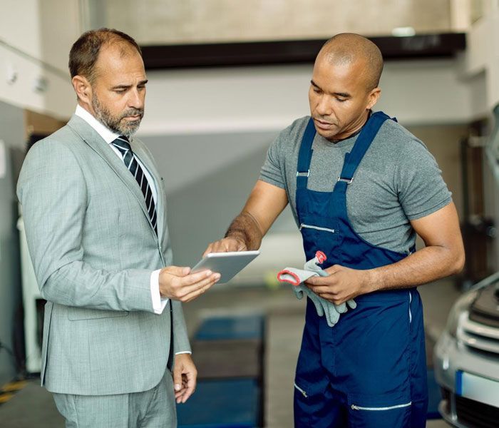 A man in a suit and tie is looking at a tablet with a mechanic.