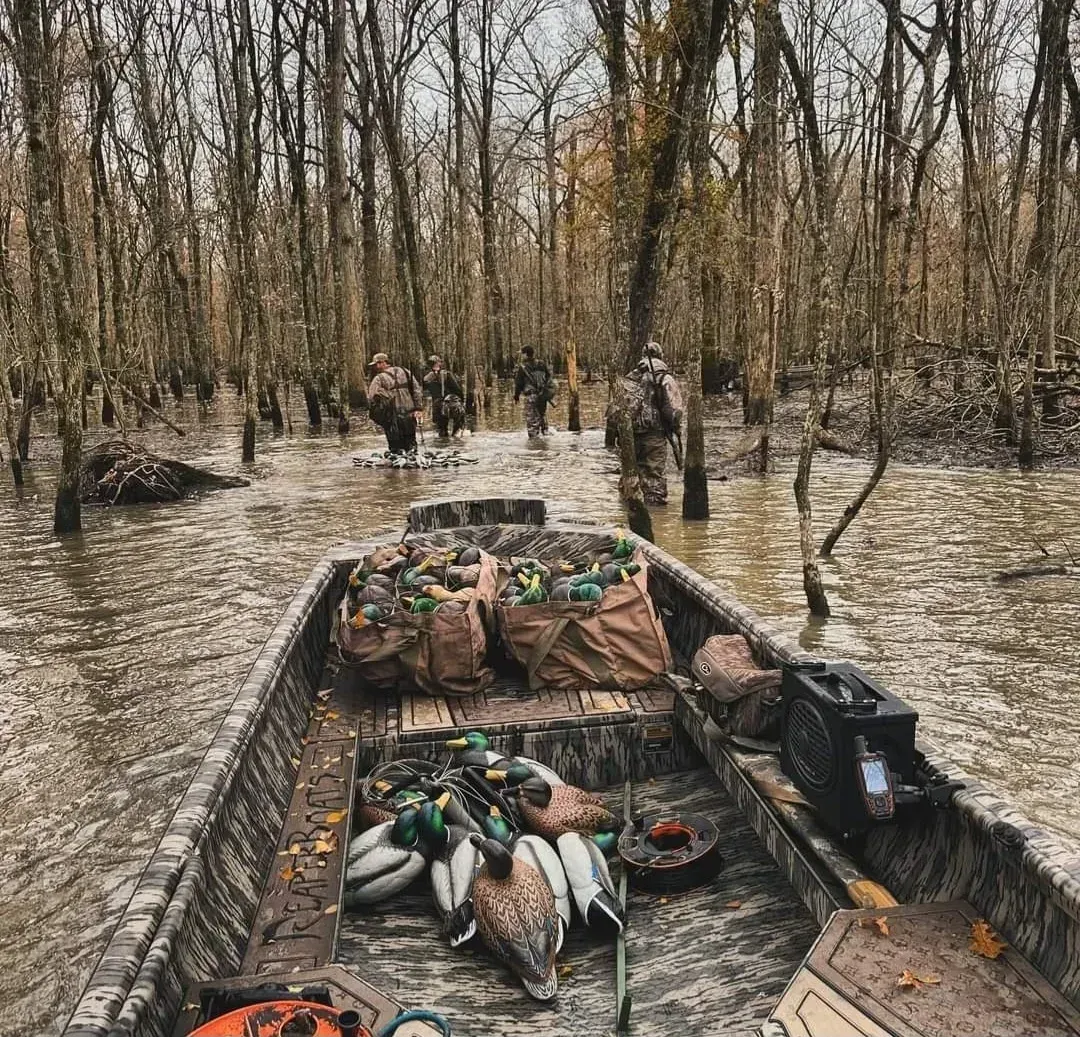 Small boat loaded with ducks and gear in a muddy flooded forest, with people wading ahead