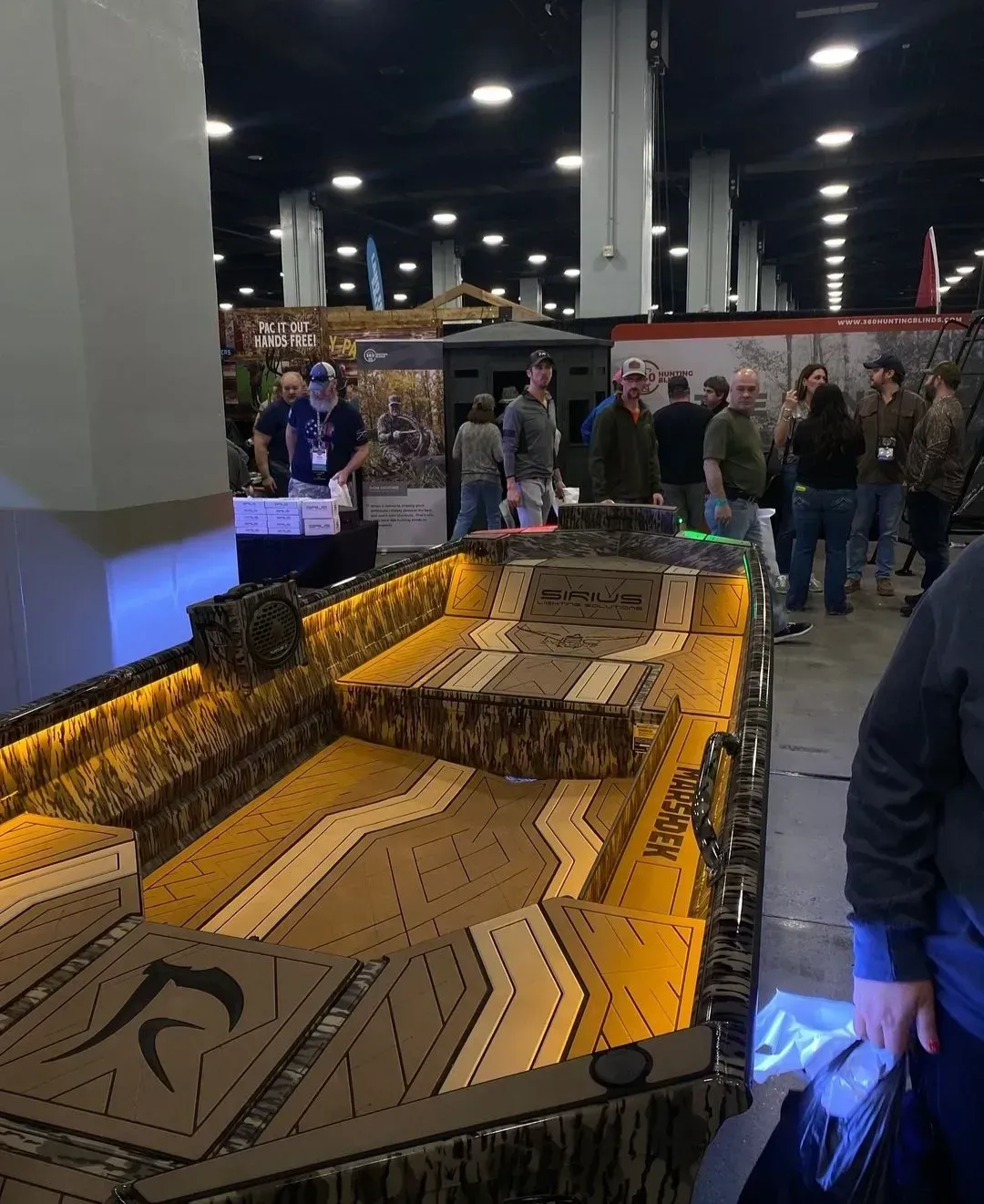 Indoor trade show display of a large geometric model with tan and blue lighting, surrounded by attendees
