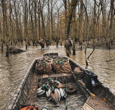 Boat loaded with supplies moving through a flooded forest, with people wading in the water nearby
