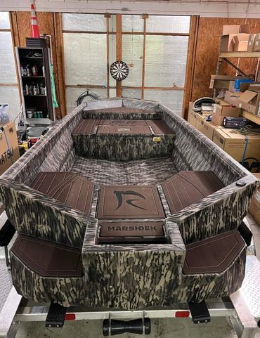Camouflaged small aluminum boat on a trailer in a workshop, with a raised central platform and flat deck.