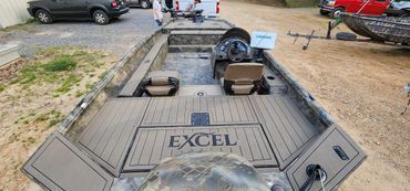 Flat-bottom boat on a trailer, with “EXCEL” on the rear deck, parked on a dirt lot near other vehicles.