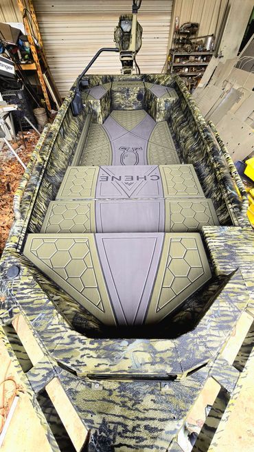 Camouflaged military-style boat indoors, viewed from above, with tan deck panels and gray seating.