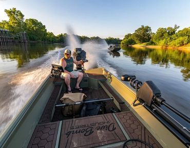 Two people fishing in a boat on a calm river at sunset, with a spray from the motor behind them