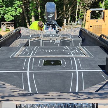 Black boat deck with fishfinder, person in neon jacket, and yellow excavator in a wooded area