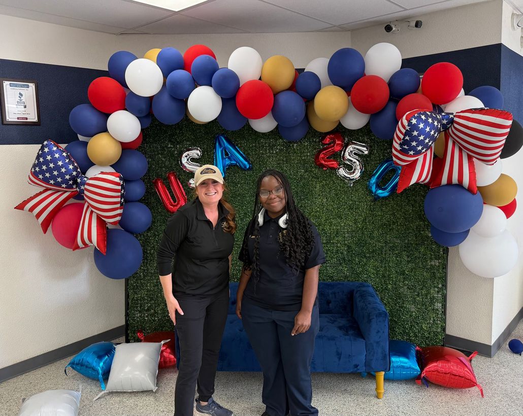 Two people posing before a red, white, and blue balloon arch and “USA 25” backdrop on a couch.
