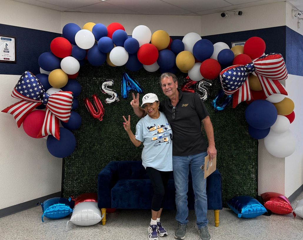 Two people posing under red, white, and blue balloons and stars, with “USA” letters on a green backdrop.