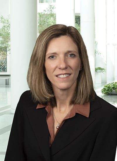 Professional headshot of a woman in a black blazer and brown blouse, smiling in an office setting