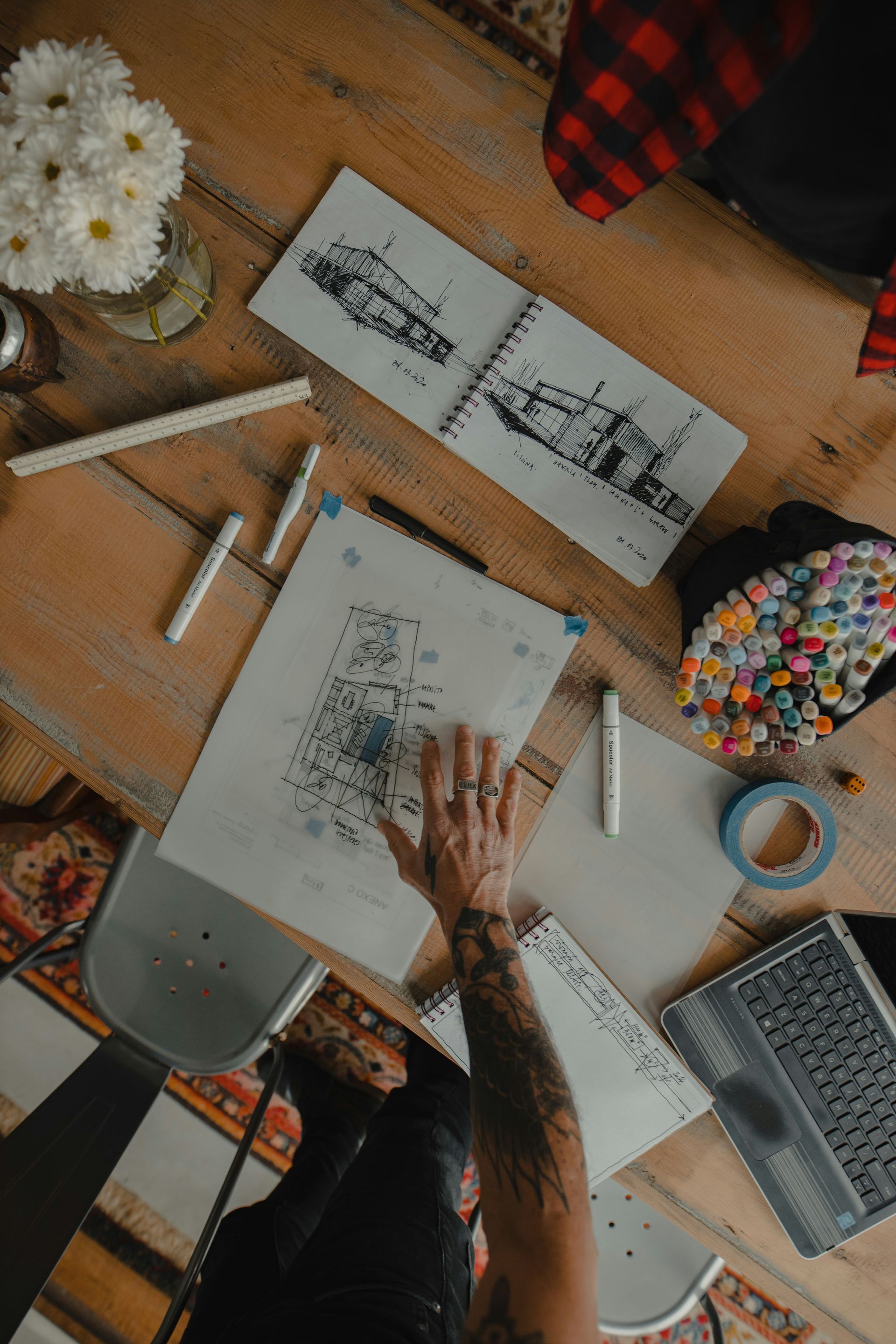 Overhead view of a desk with sketchbooks, hands drawing, flowers, pencils, laptop, and coffee cup