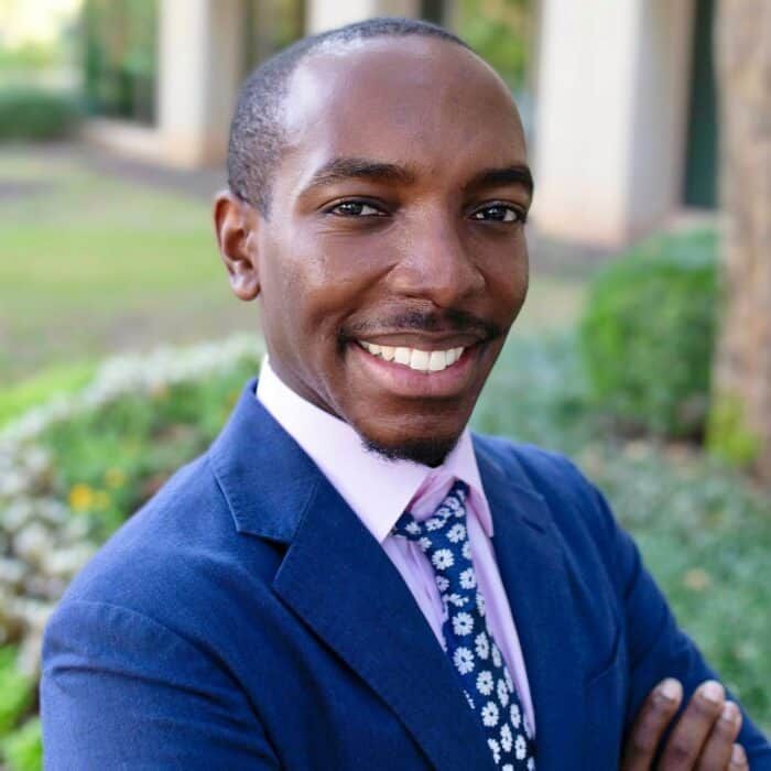 Smiling man in a blue suit and patterned tie outdoors with a blurred garden background