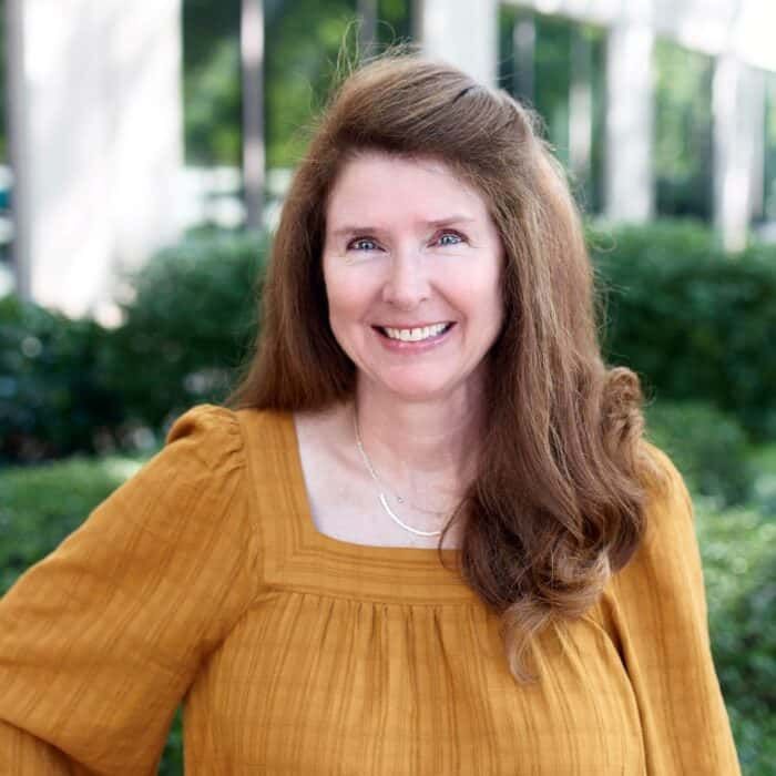Smiling woman in a mustard blouse outdoors, with greenery and building columns behind her