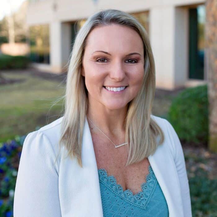 Smiling woman in white blazer and blue top standing outdoors near a building