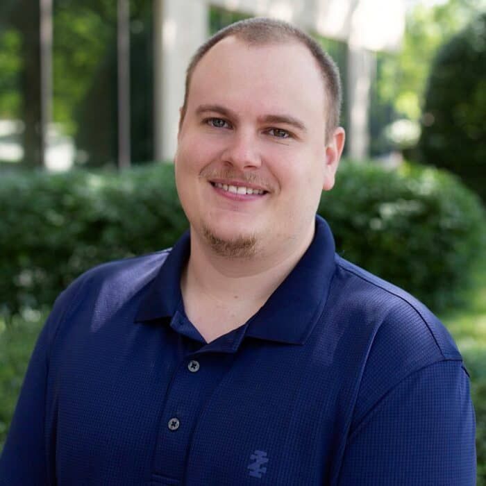 Smiling man in a navy polo shirt standing outdoors with blurred greenery behind him