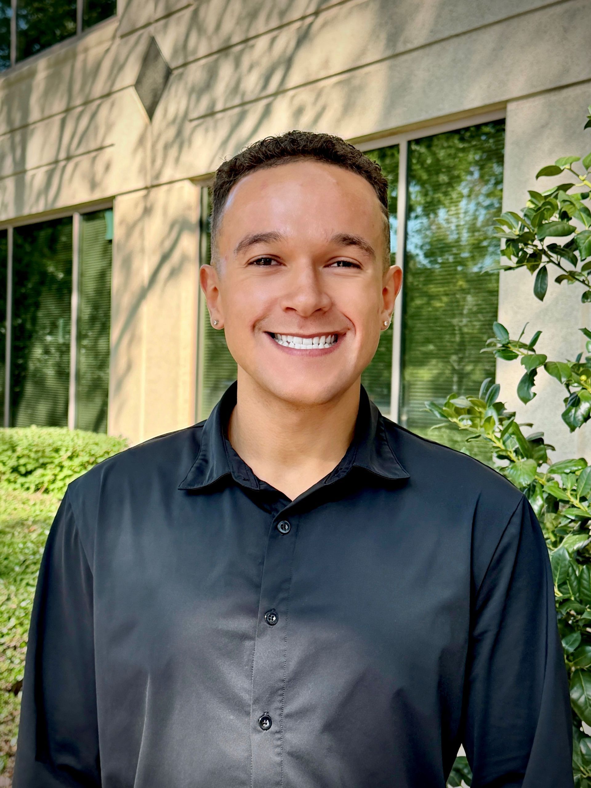 Smiling person in a dark shirt standing outdoors beside a building with glass windows and greenery