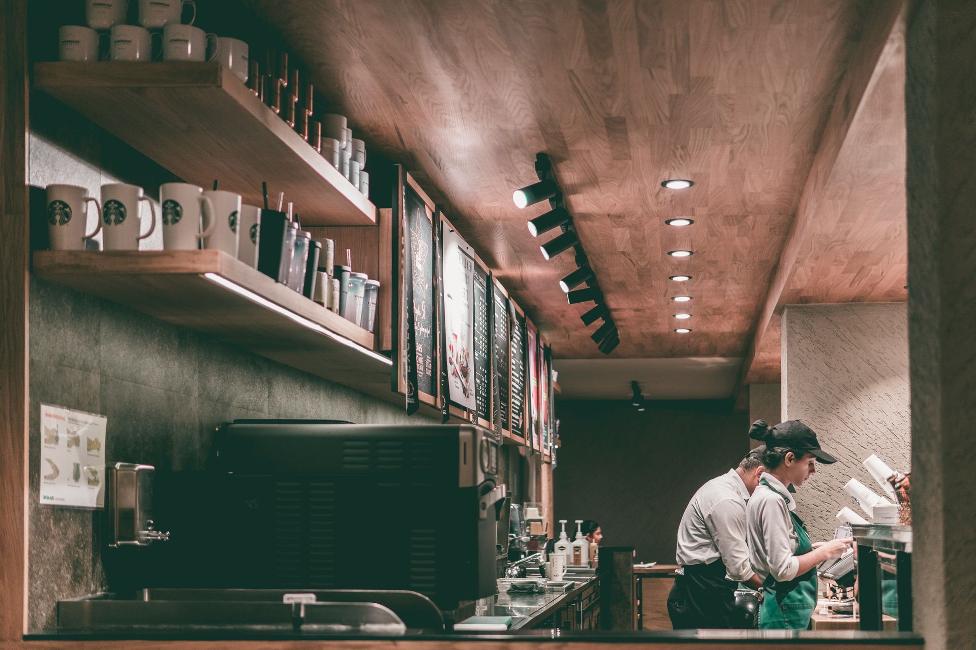 Two baristas working behind the counter in a cozy coffee shop with warm wood shelves and hanging lights