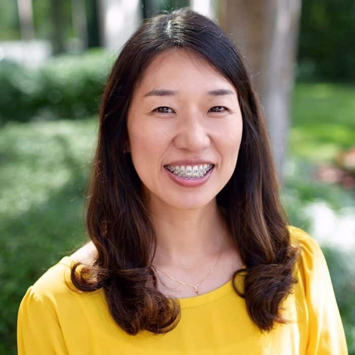 Smiling person with long dark hair in a yellow top outdoors, with a blurred green background