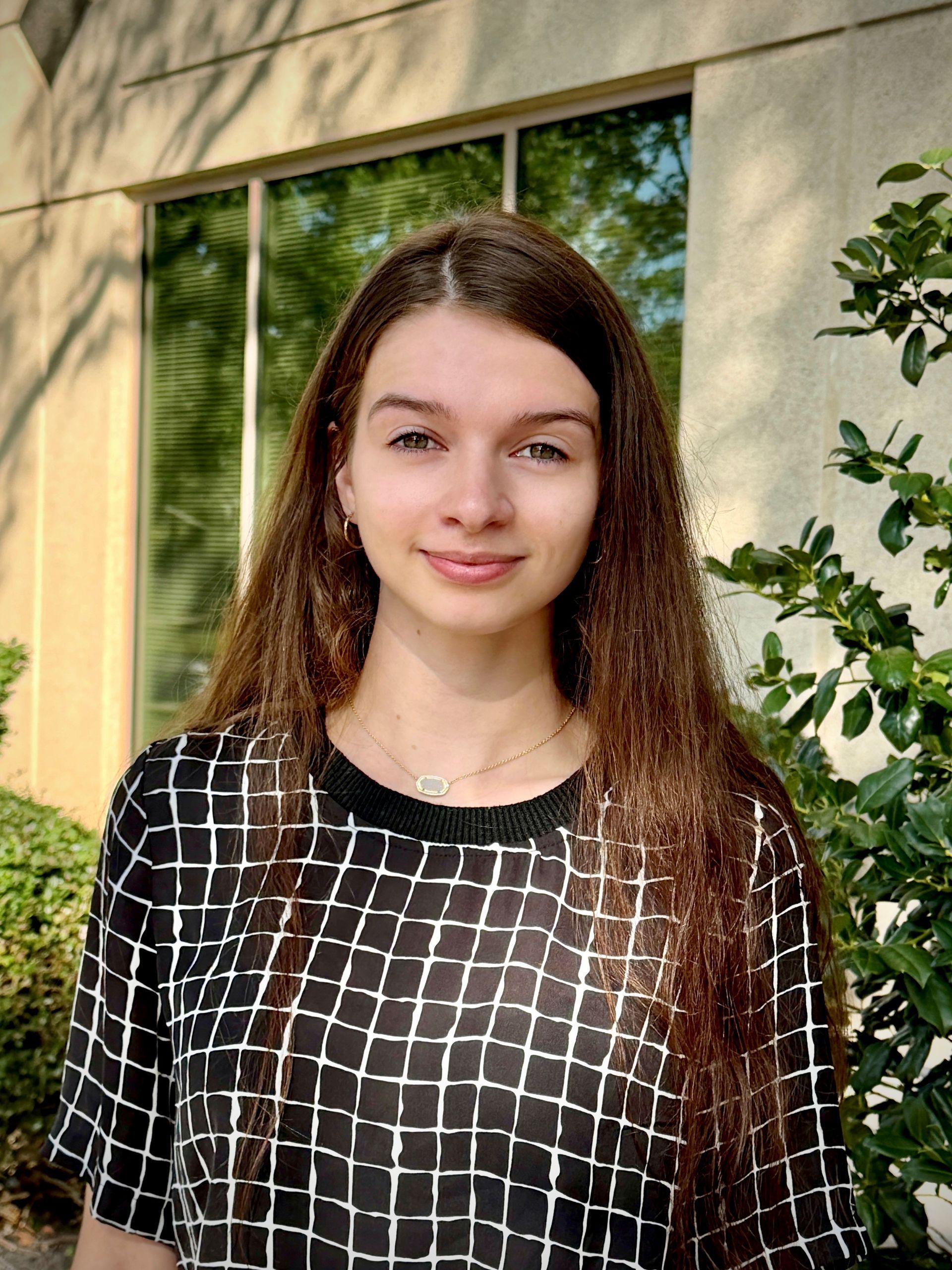 Young person standing outdoors by a beige wall and green plants, wearing a black-and-white checkered shirt.