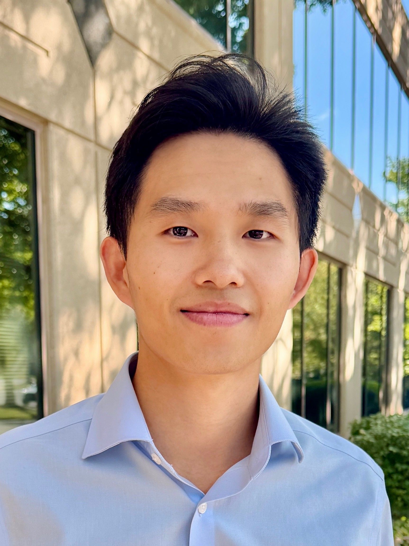 Portrait of a person in a light blue shirt standing outdoors by a building with windows and greenery.