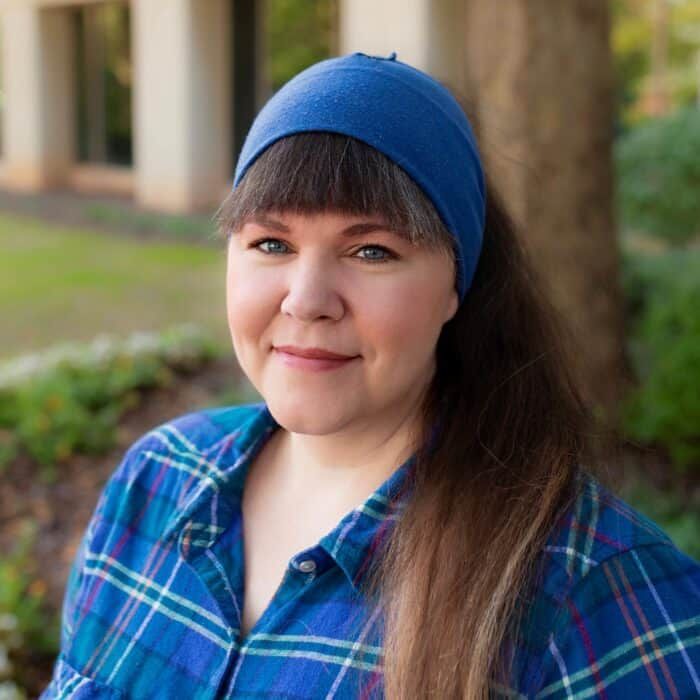 Portrait of a smiling person in a blue headband and plaid shirt outdoors, with trees softly blurred behind.