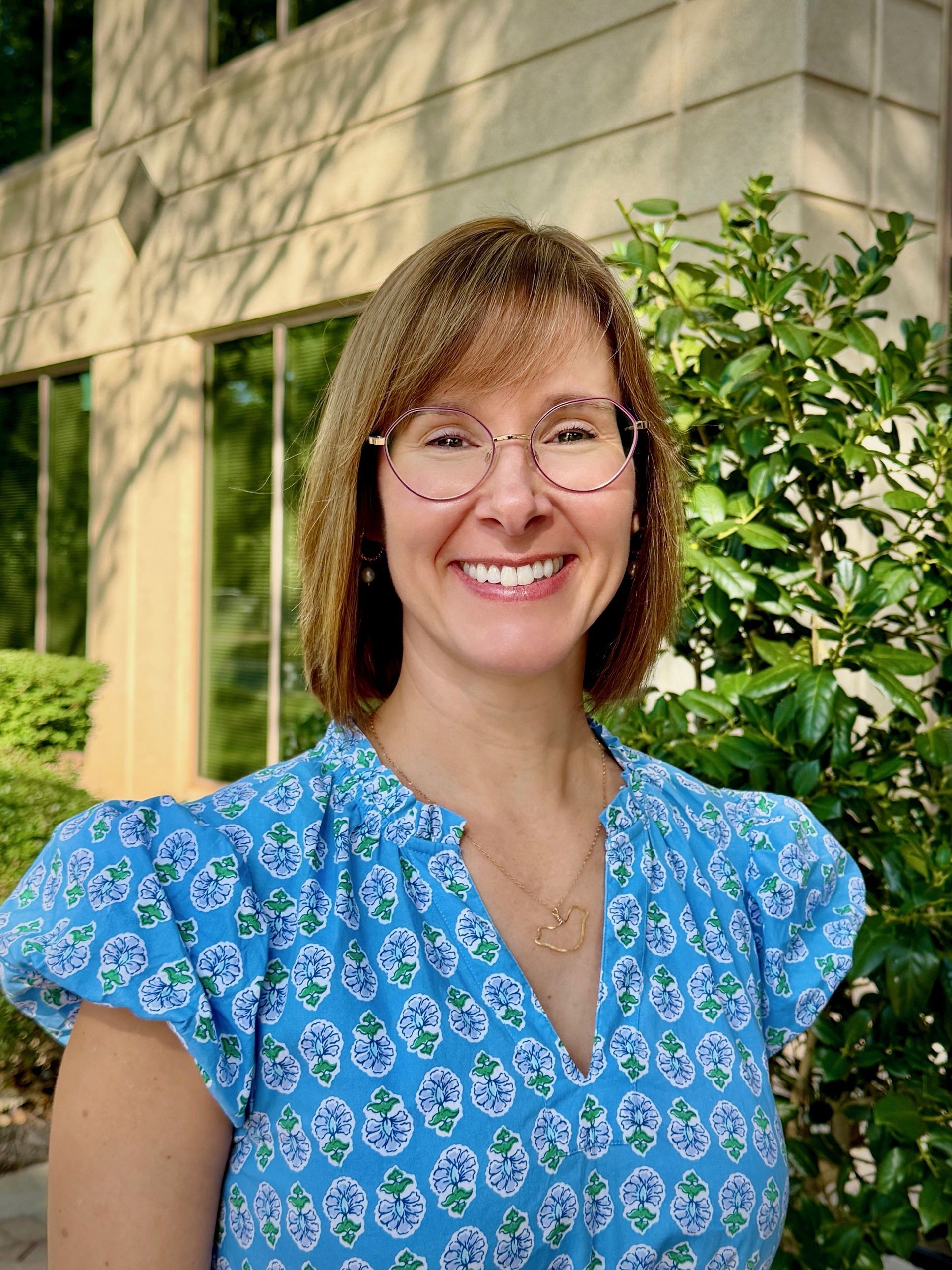 Smiling person with short hair and glasses wearing a white blouse outdoors.
