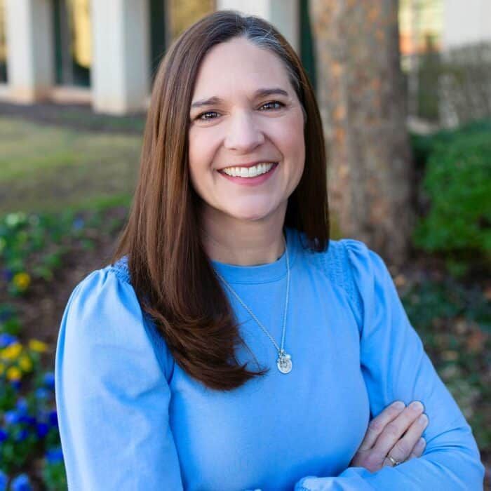 Smiling woman in a blue top standing outdoors with arms crossed, near trees and a building.