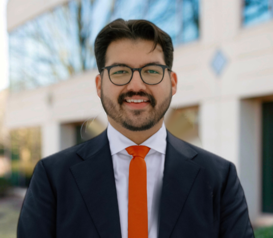 Smiling man in glasses and navy suit with orange tie outdoors in front of a building