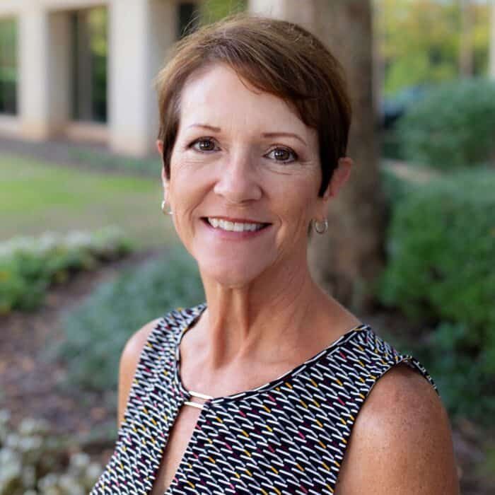 Smiling woman standing outdoors near trees and shrubs, wearing a patterned sleeveless top