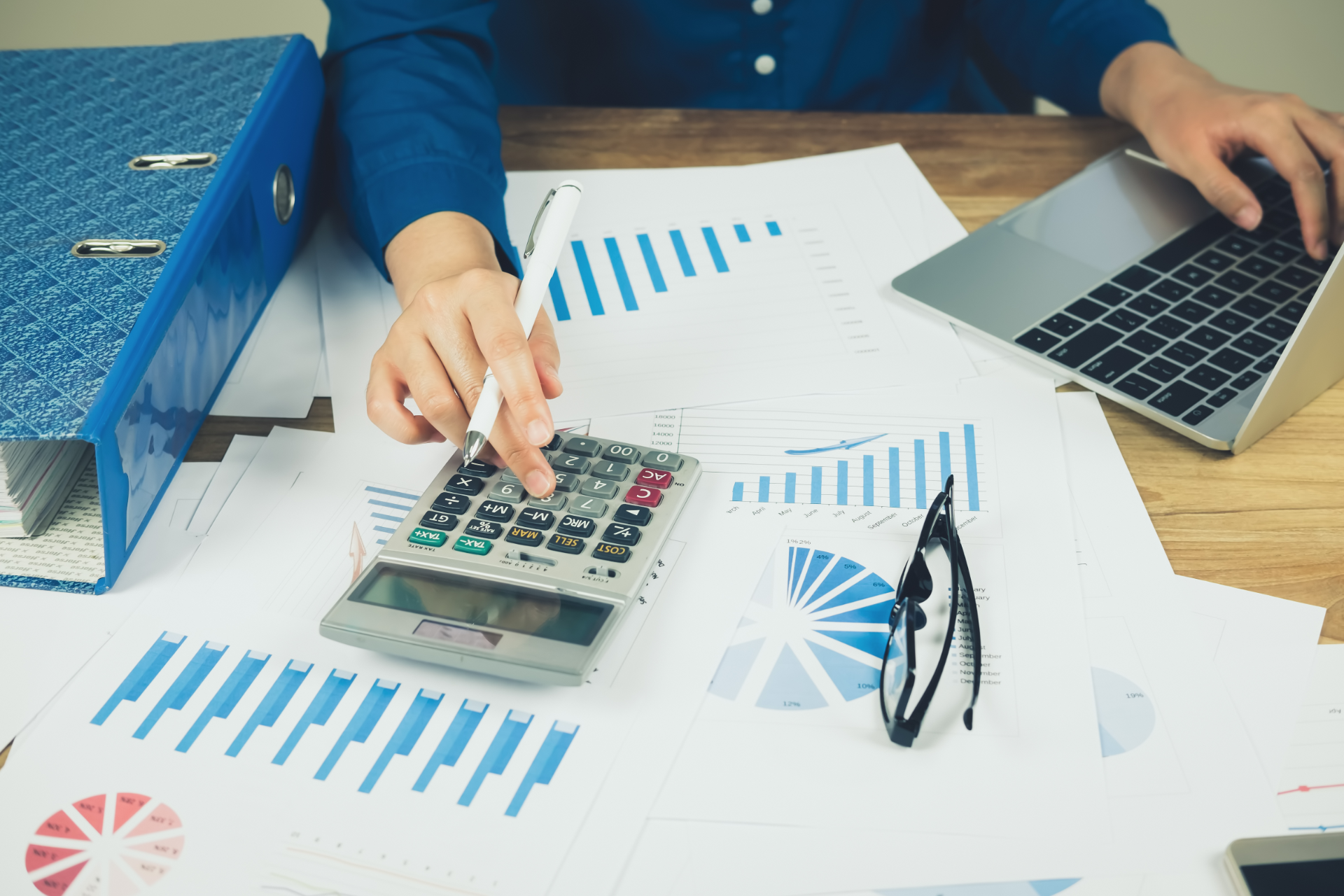 Person using a calculator over financial charts beside a laptop and eyeglasses on a desk