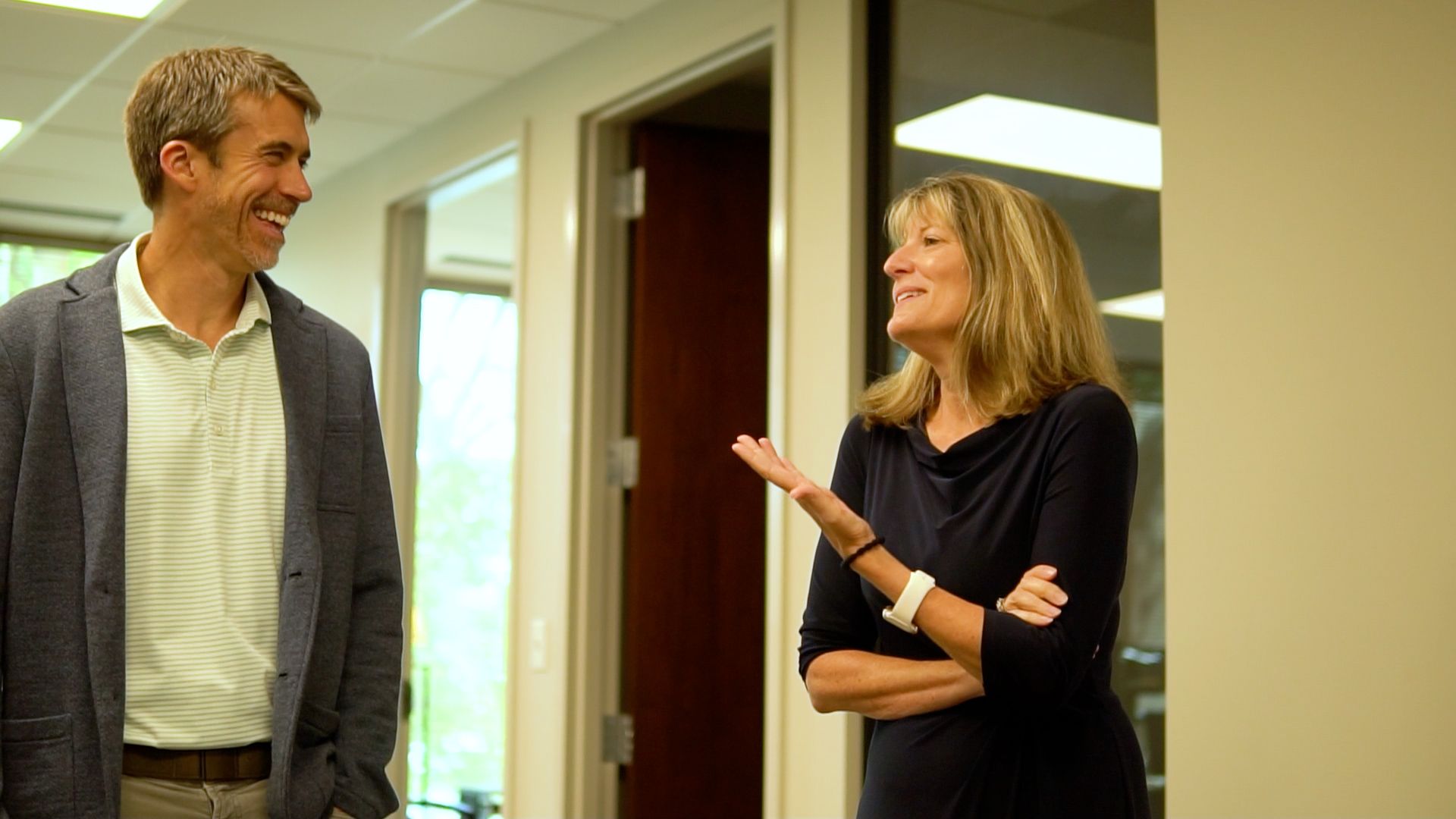 Two people talking in a hallway, one gesturing while the other smiles.