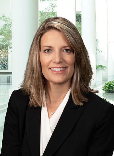 Professional headshot of a smiling person in a black blazer and white blouse against a bright office background