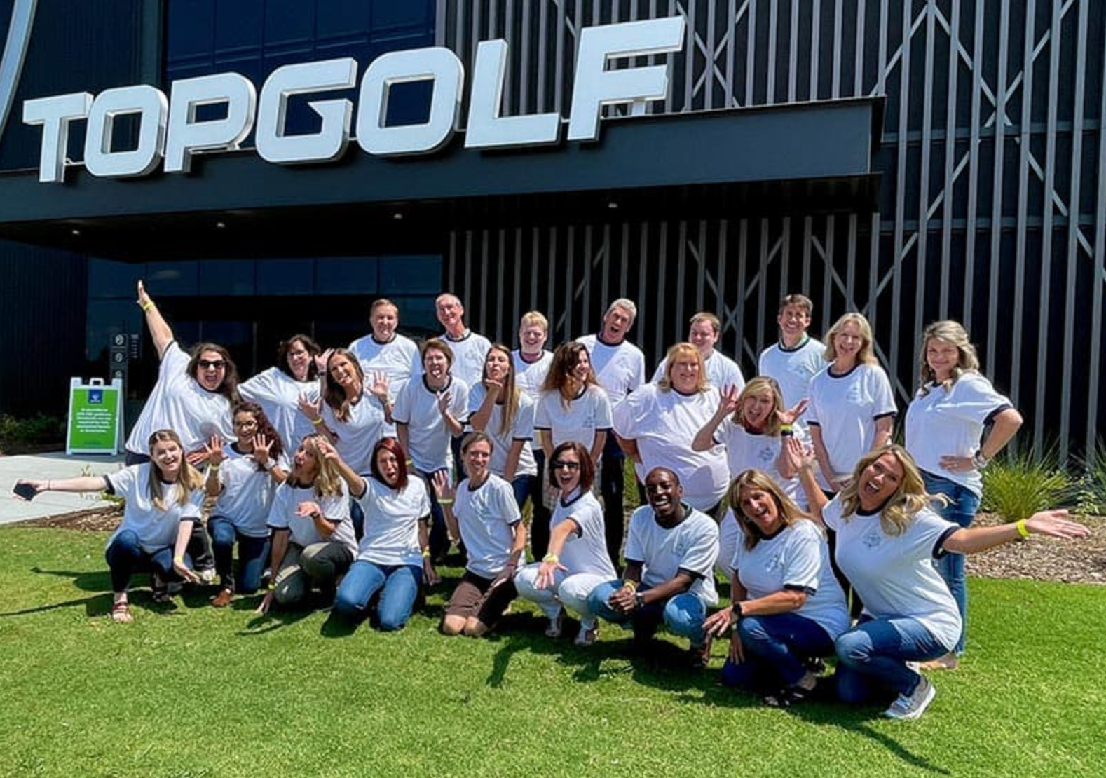 Group of people posing outside Topgolf, many in white shirts, smiling on a sunny day