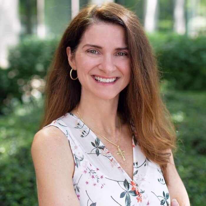 Smiling woman with long brown hair in a white floral sleeveless top outdoors, with greenery behind her