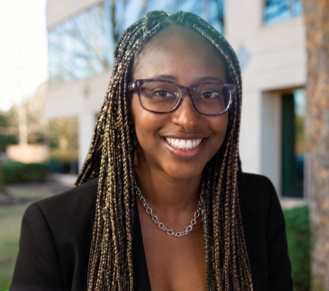 Smiling woman with glasses and braided hair standing outdoors in a black blazer