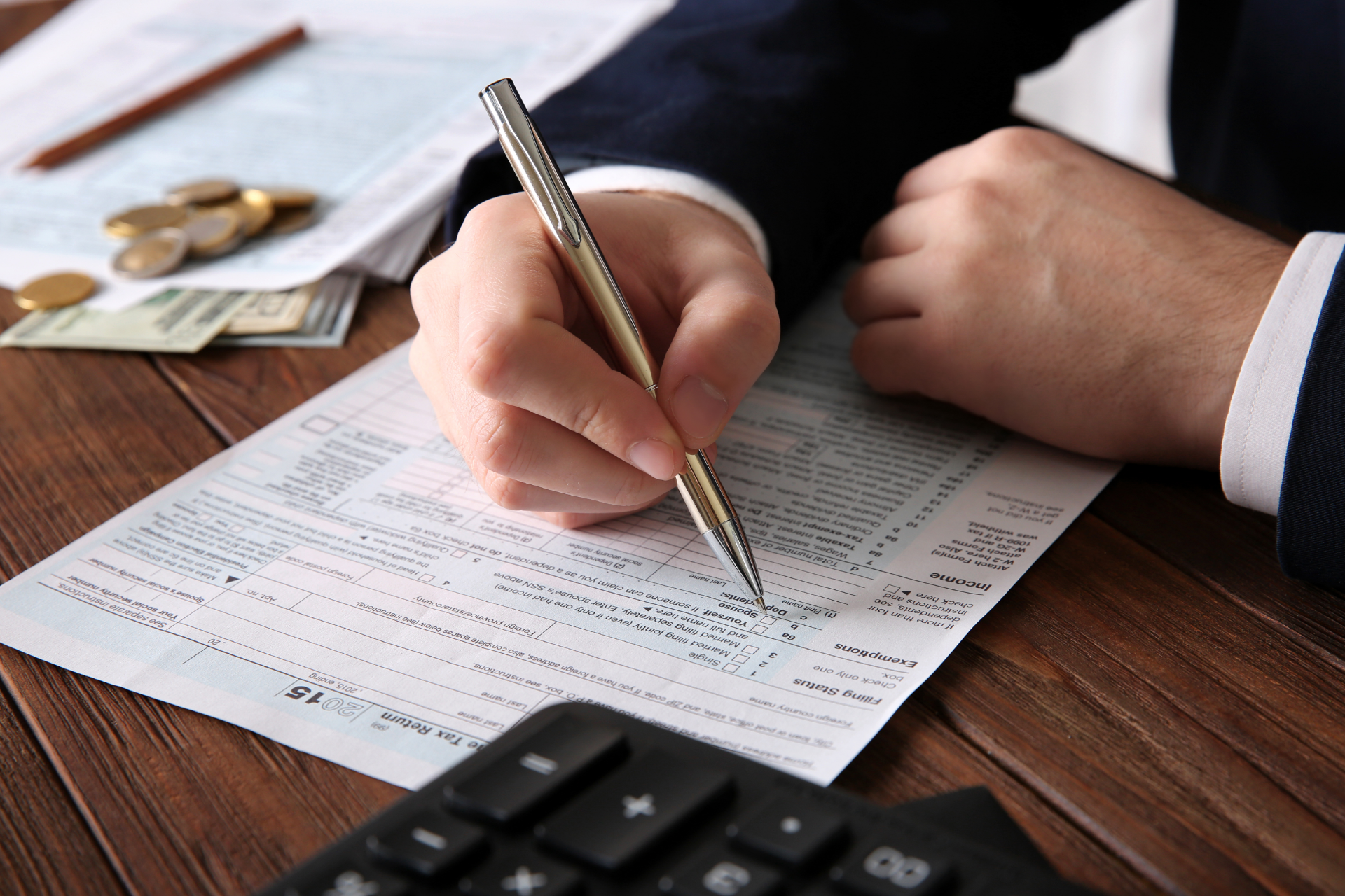 Person signing a document with a pen on a wooden desk, with coins, bills, and a calculator nearby