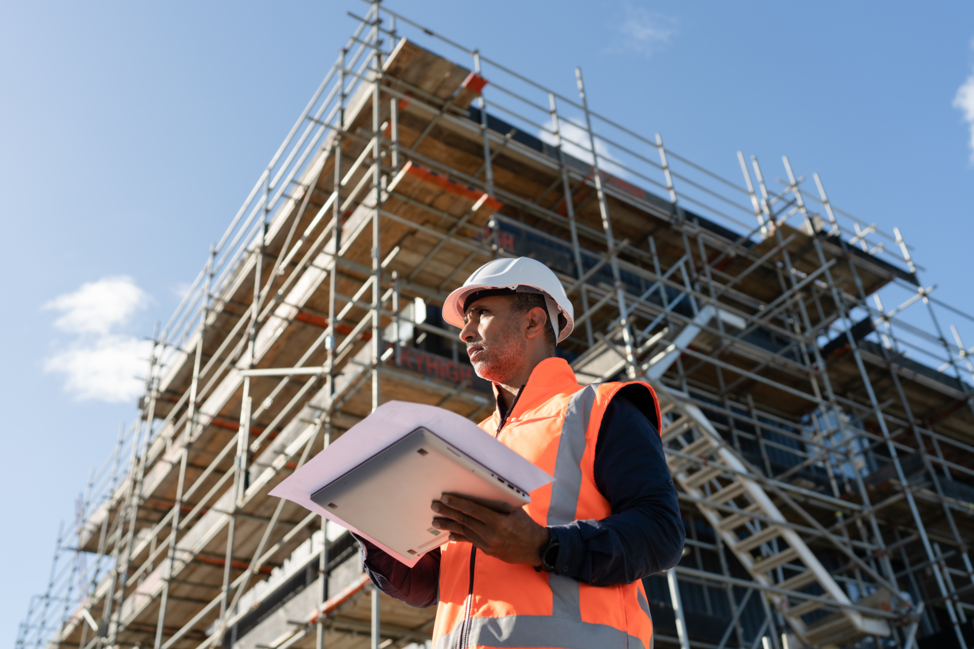 Worker in orange vest and hard hat using a tablet at a construction site with scaffolding.
