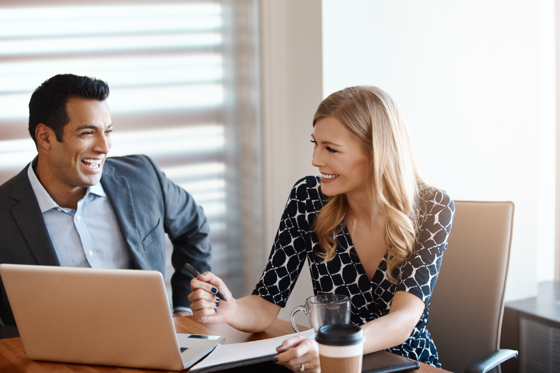 Two coworkers smiling and talking at a desk with a laptop in a bright office