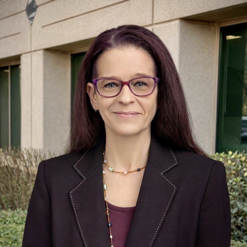 Woman in glasses and dark blazer standing outdoors in front of a building.